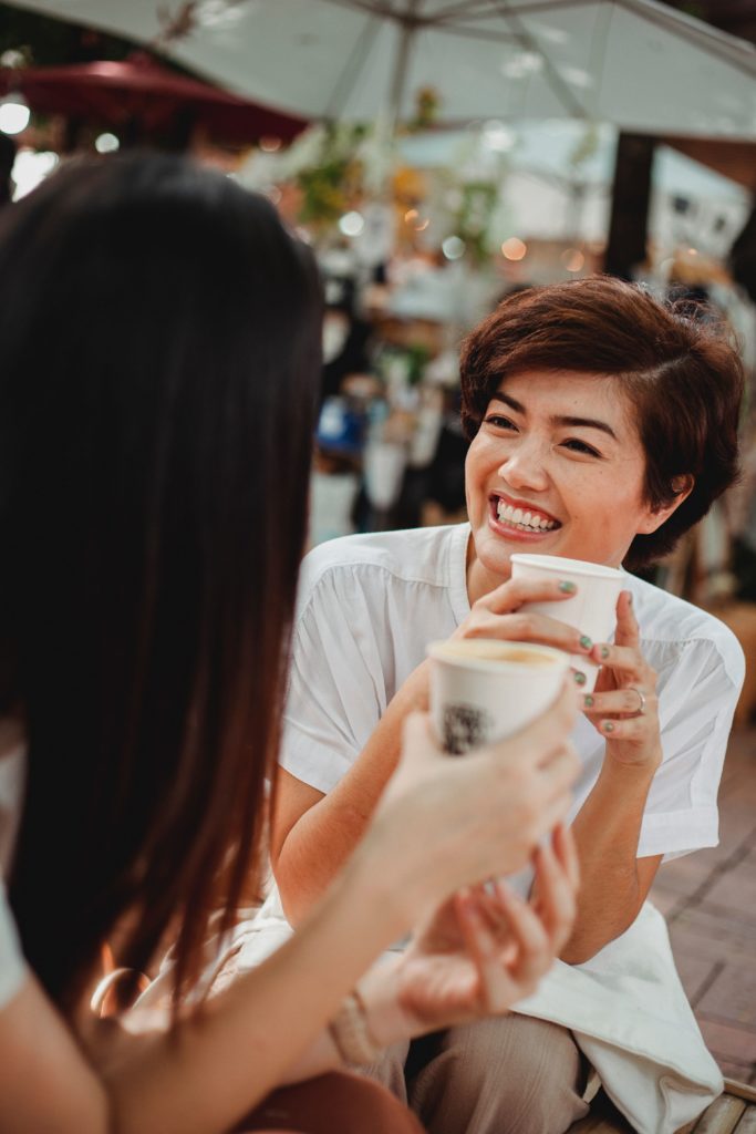 une femme qui fait fasse à une autre durant sa pause, toutes les deux ont un gobelet de café à la main et sourient