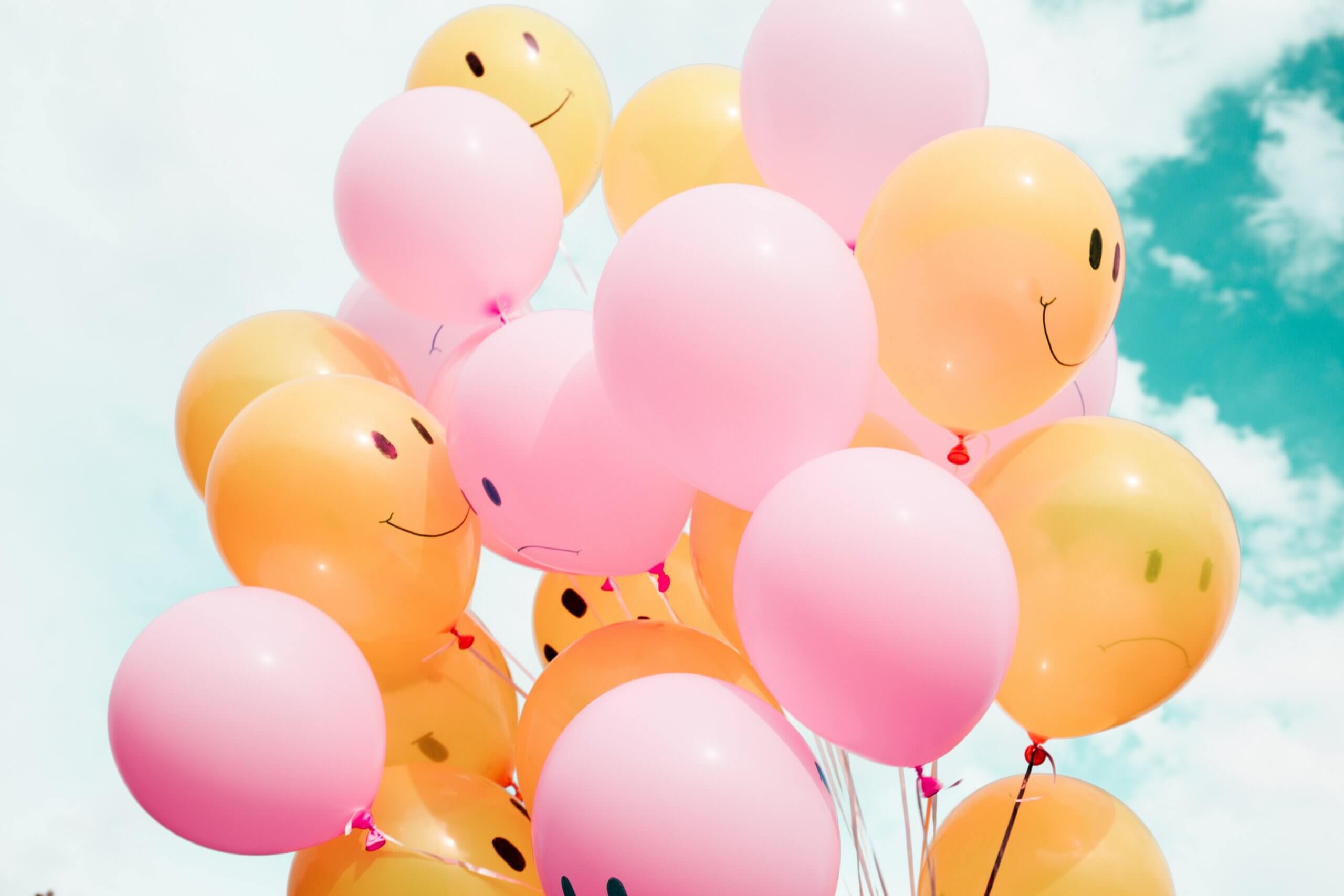 Plusieurs ballons de couleur rose et orange avec un sourire dessiné dessus, dans le ciel